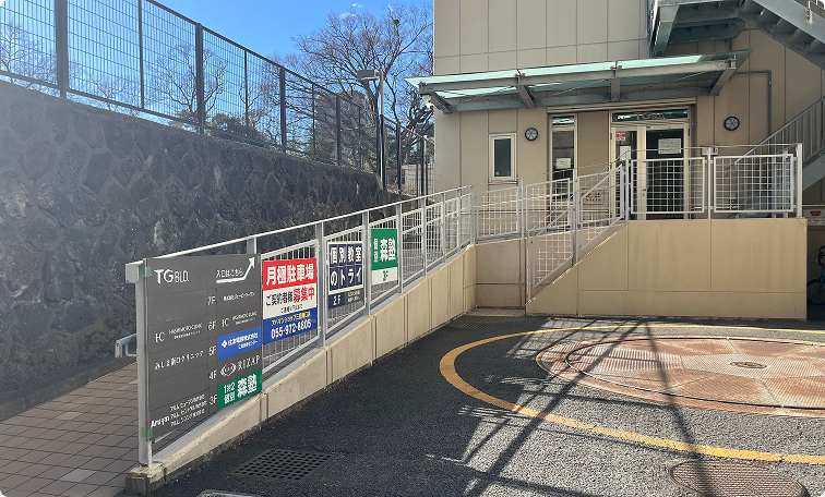 細い道に入るとすぐに｢TGビル｣と書かれた青い案内板があります。 その案内板の後ろにあるスロープを上がって、ビルの入り口へお進みください。